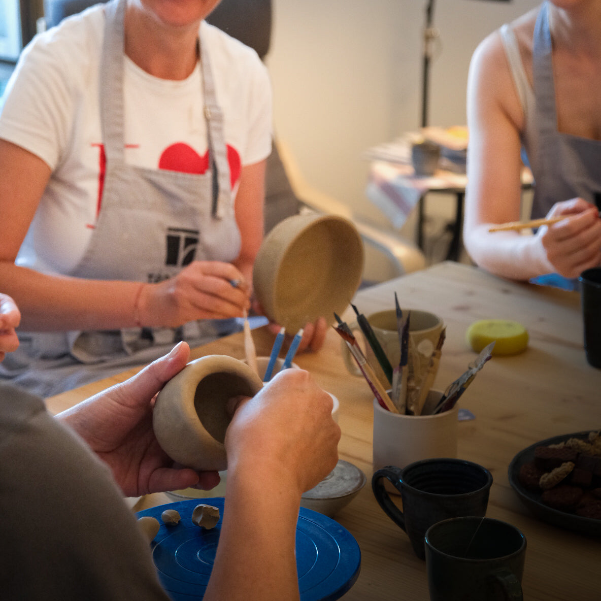 Im Tonraum Kaffeekränzchen modellieren und bemalen drei Personen gemeinsam Tonschalen. Umgeben von Malutensilien, Kaffeetassen und Pinseln töpfern sie als Team - eine Person hält eine Schale, eine andere verziert sie, alle tragen Schürzen.