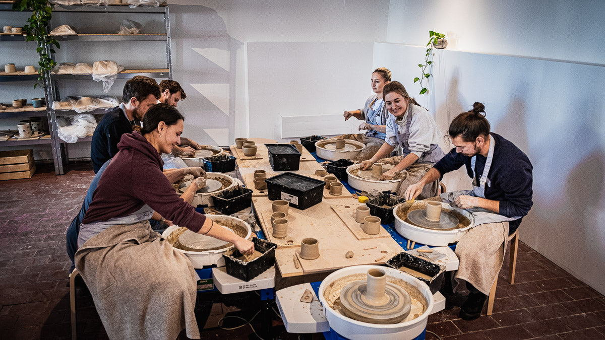 Eine Gruppe von Menschen sitzt lächelnd um einen Tisch herum und formt Ton auf Töpferscheiben in einem hellen Studio mit Regalen voller Töpferwaren im Hintergrund.