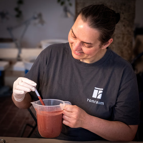 Mit Handschuhen und einem Tonraumhemd rührt jemand lächelnd eine rötliche Flüssigkeit in einem Krug um - eine Szene aus unserem Kurs "Glasuren im Griff - Tauchglasuren erfolgreich anwenden". Der sanft verschwommene Hintergrund unterstreicht den kreativen Prozess.