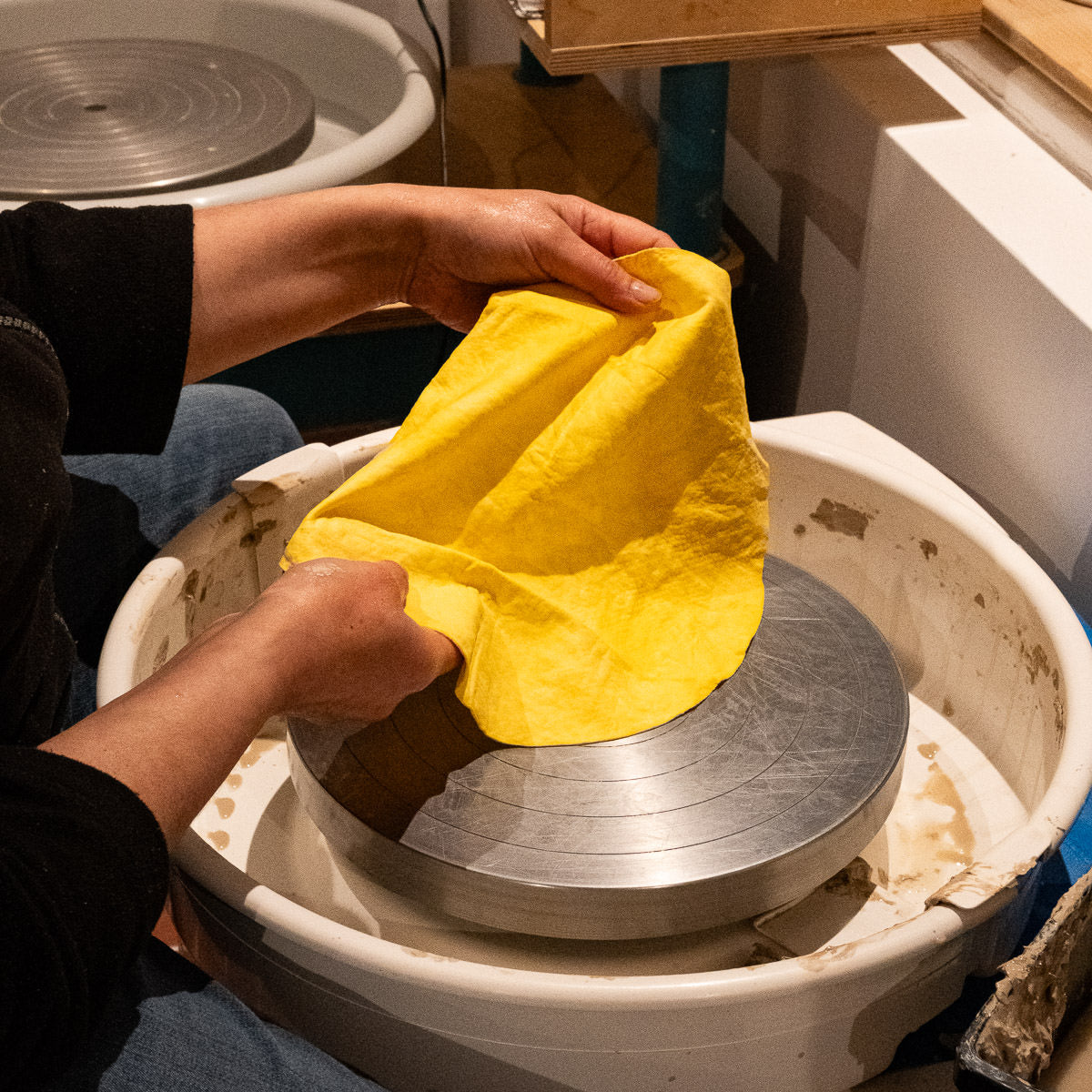 Eine Person bedeckt eine Drehscheibe mit einem leuchtend gelben Bat Mate und bereitet sich auf die Arbeit in einer Keramikwerkstatt vor. Ihre Hände halten die Bat Mate über dem Kopf der Metalldrehscheibe, wobei Tonreste um die Schale herum sichtbar sind.