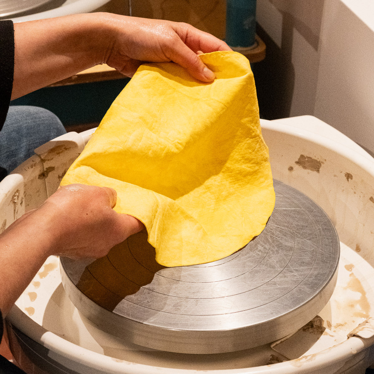 Die Hände einer Person legen einen leuchtend gelben Bat Mate über eine saubere, silberne Töpferscheibe und bereiten sich auf die Arbeit in einem Atelier vor, in dem im Hintergrund Geräte zu sehen sind.