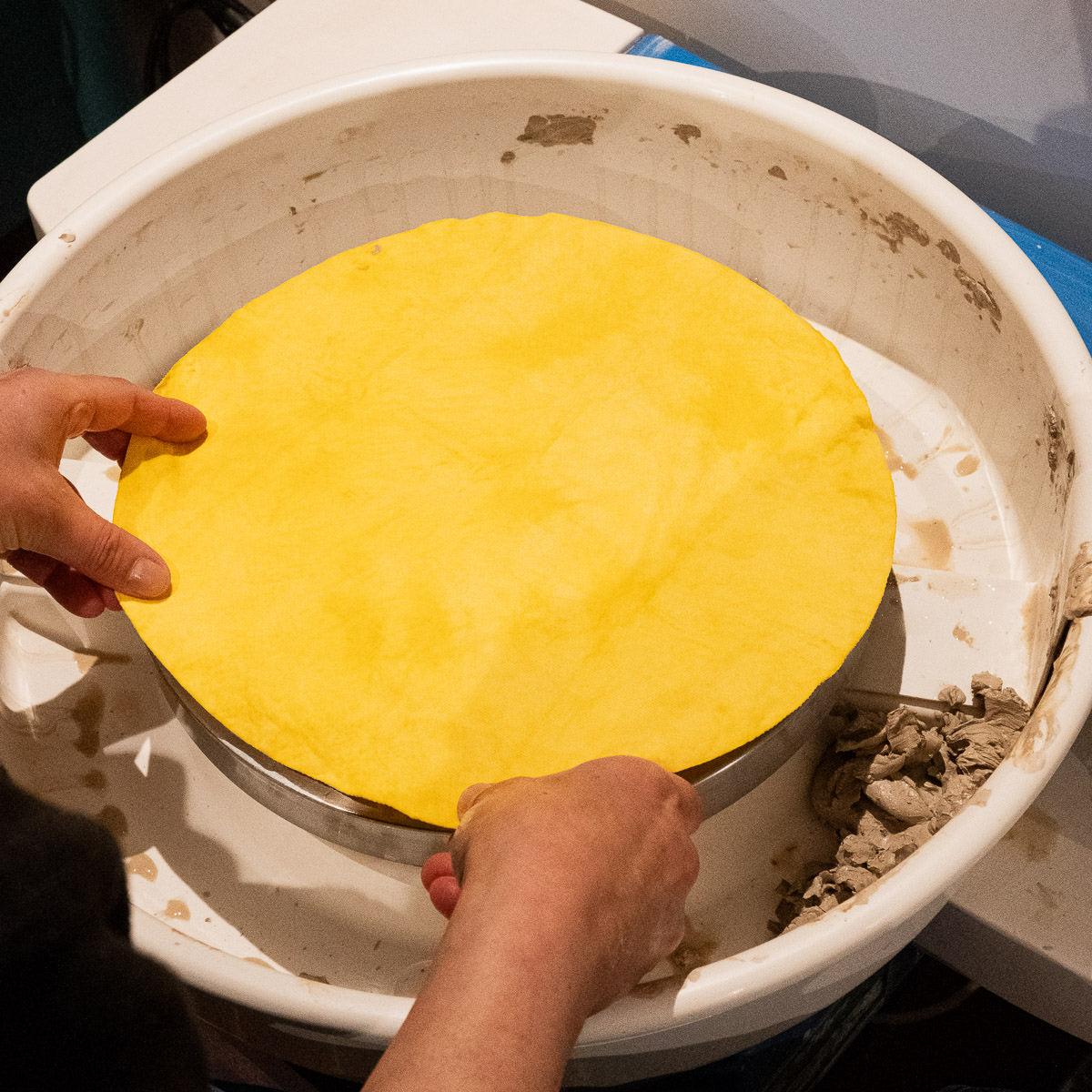 Eine Person legt ein rundes, gelbes Teigblatt auf den Bat Mate in einem weißen Behälter und formt es vorsichtig mit beiden Händen.