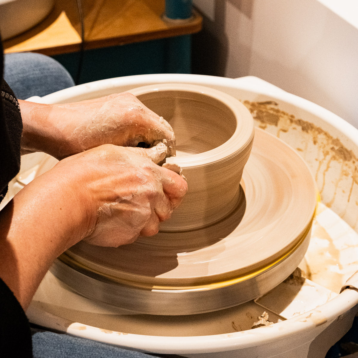 Nahaufnahme der Hände einer Person, die eine Tonschale auf dem Bat Mate formt, wobei um den Arbeitsbereich herum nasser Ton, Werkzeuge und ein Reinigungstuch zu sehen sind.