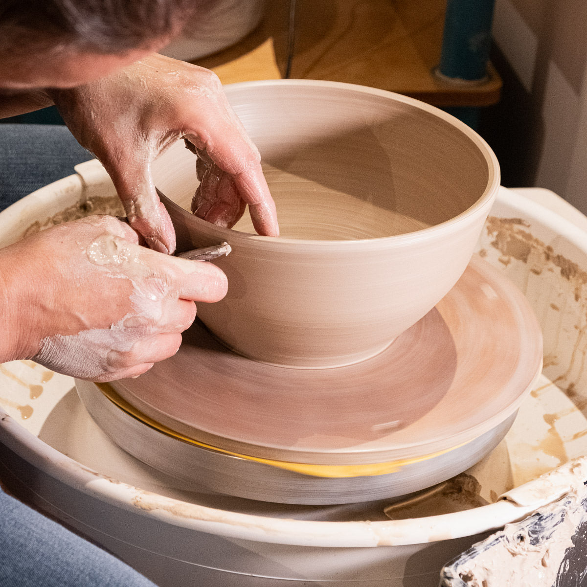 Eine Person formt eine Tonschale auf einem Bat Mate, wobei sie beide Hände benutzt, um den sich drehenden Ton zu formen, mit Töpferwerkzeugen, einem Reinigungstuch und nassem Ton in der Nähe.