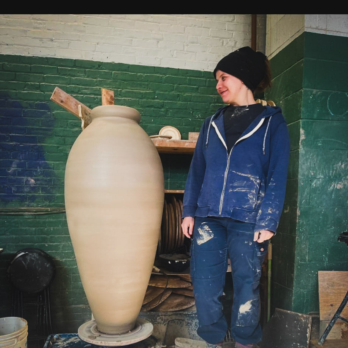 Eine Frau in mit Farbe bespritzter Kleidung und einer Mütze lächelt neben einer großen, unvollendeten Tonvase in einer Werkstatt mit grünen und weißen Backsteinwänden während Einmalig: Großformat-Drehen mit Eva Dürnegger.