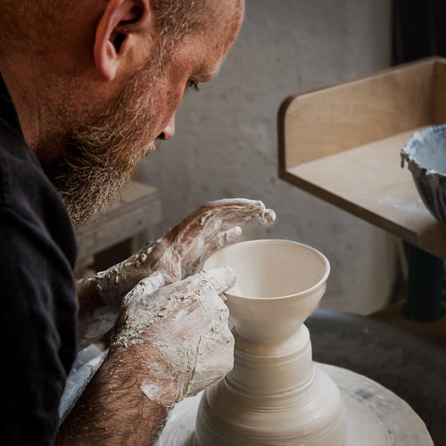 Ein bärtiger Mann formt in einem Atelier einen Tontopf auf einer Drehscheibe, seine Hände sind mit nassem Ton bedeckt, während er sich konzentriert - ein Beispiel aus der "Masterclass": Serien drehen.