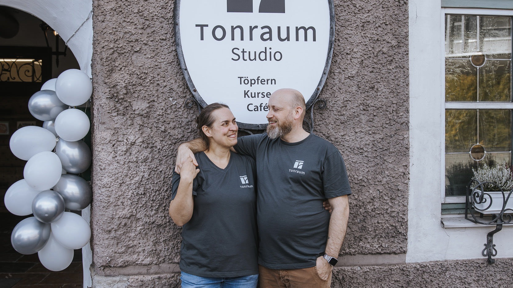 Zwei Menschen in passenden Tonraum Studio-Shirts stehen lächelnd und mit umschlungenen Armen vor einem Gebäude mit einem Tonraum Studio-Schild, neben einem Haufen weißer und silberner Luftballons.