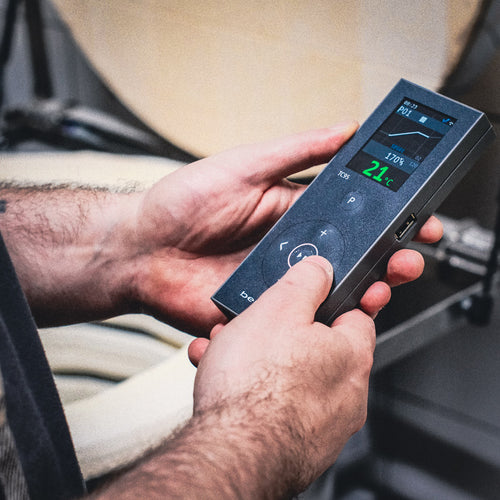 Eine Person hält eine rechteckige Fernbedienung für "Besser brennen im Elektro-Ofen - Brennkurven einfach erklärt", deren digitaler Bildschirm 21°C und Einstellungen anzeigt. Ihre Arme sind vor einem leicht verschwommenen Hintergrund zu sehen.