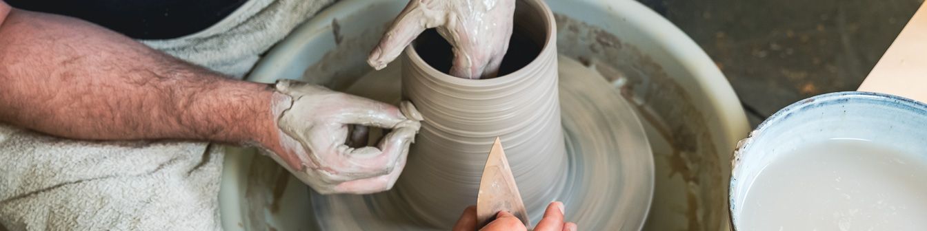 Zwei Hände formen einen Tontopf auf einer rotierenden Töpferscheibe, während eine andere Hand die Oberfläche mit einem Holzwerkzeug glättet. Ein Eimer mit Wasser steht daneben, und über einem Arm hängt ein Handtuch.